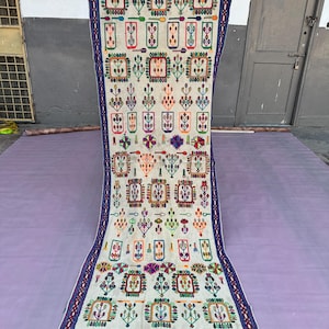 May include: A long, rectangular, off-white rug with colorful embroidered floral and geometric patterns. The rug has a blue border with red accents and a fringe along the bottom edge. The design includes squares, rectangles, and stylized plant motifs.
