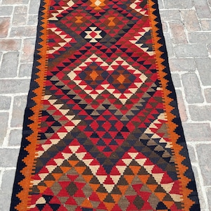 Puede incluir: Una alfombra rectangular tejida a mano con un patrón geométrico. La alfombra presenta un diseño central de diamantes en rojo, marrón y crema, enmarcado por un borde naranja y negro. Las dimensiones de la alfombra no son visibles.