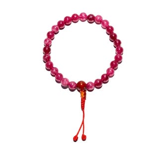 May include: A beaded bracelet featuring small, round, pink and red beads. A larger, reddish-brown bead is centered, with a red tassel hanging below. The bracelet is set against a white background.
