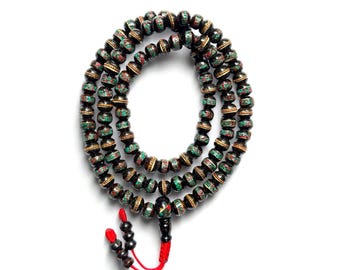 Puede incluir: Un collar de cuentas redondas en negro, dorado, rojo y verde. Las cuentas están ensartadas juntas, con una borla roja en el extremo. El collar se muestra sobre un fondo blanco.