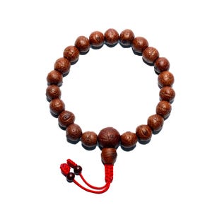 Puede incluir: Una pulsera de cuentas marrones con borla y detalles rojos. La pulsera presenta múltiples cuentas redondas y una cuenta central más grande. La borla es roja con dos pequeñas cuentas marrones oscuras. Sobre fondo blanco.