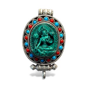 Pode incluir: Pingente oval com uma escultura de divindade turquesa, emoldurado por uma borda prateada. A borda é adornada com pedras preciosas vermelhas e azuis alternadas. O pingente tem uma argola prateada para pendurar.