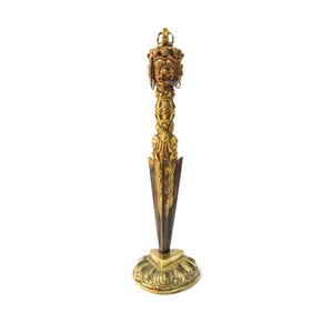 May include: A gold-colored, ornate dagger with a detailed handle featuring a face and crown design. The blade is a contrasting bronze color, and the dagger rests on a decorative, circular base. The overall design suggests a ceremonial or decorative object.