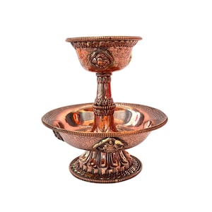 Puede incluir: Un cuenco decorativo de dos niveles de color cobre con intrincados patrones florales y geométricos. El cuenco superior es más pequeño y se asienta sobre un pedestal detallado, mientras que el cuenco inferior es más grande y ancho. La pieza tiene un acabado pulido.