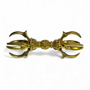 Op de afbeelding: Een goudkleurige vajra, een ritueel object met een centrale bol en drie tanden die zich van elk uiteinde uitstrekken. Het object heeft ingewikkelde details en wordt tegen een witte achtergrond getoond.