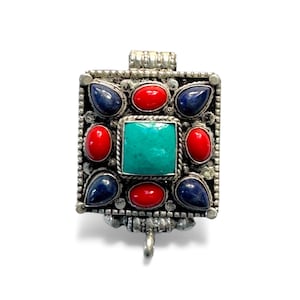 May include: A square silver pendant with a central turquoise stone, surrounded by red and blue gemstones. The pendant has a decorative border and a small loop at the bottom for attachment. The design is symmetrical and ornate.