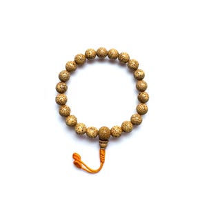 May include: A bracelet made of light brown, speckled beads. It features an orange tassel. The beads are arranged in a circular pattern against a white background. The beads are approximately 1 cm in diameter.