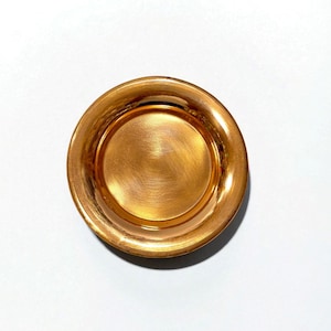 May include: A top-down view of a small, round, copper-colored bowl. The bowl has a wide, slightly sloped rim and a smaller, recessed center. The surface has a brushed metal texture, reflecting light.