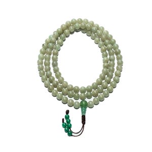 Puede incluir: Un collar de cuentas de jade verde claro, dispuestas en tres bucles, con un colgante de jade verde y una borla marrón. Las cuentas son redondas y lisas, y el colgante está tallado. El fondo es blanco.