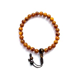 Puede incluir: Una pulsera de cuentas de madera marrón claro con vetas marrón oscuro. La pulsera tiene una borla marrón oscuro y una sola cuenta más grande, marrón oscuro. La pulsera se muestra sobre un fondo blanco.