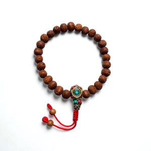 May include: A brown beaded bracelet with a decorative centerpiece featuring turquoise and red accents. The bracelet is strung on a red cord with wooden beads at the end. The beads are approximately 1.3 cm in diameter.