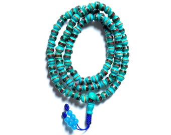 May include: Turquoise beaded necklace with red and silver accents. The beads are cylindrical and strung together, forming multiple strands. A blue tassel hangs from the bottom. The necklace is displayed on a white background.