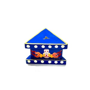 May include: A blue, house-shaped decorative box with a triangular roof and a rectangular base. The box features white dots, gold trim, and a central design with a white owl-like figure and red and orange flame-like accents. The box is painted with bright colors.
