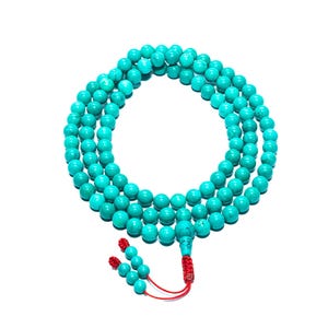 May include: A turquoise beaded necklace with a red tassel and a white background. The necklace is made of round, smooth beads. The tassel is made of small turquoise beads and red string. The necklace is likely used for meditation or as a fashion accessory.