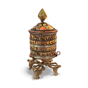 May include: A detailed, ornate prayer wheel with a conical top and a cylindrical body. The wheel is made of gold and copper-colored metal, with intricate carvings and script. It rests on a decorative, three-legged stand.