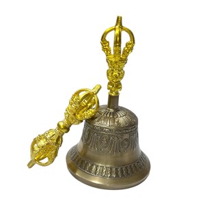 May include: A golden-colored bell with intricate patterns and a decorative handle. A matching golden hand-held bell rests beside it. The bell has a detailed design and is likely used for spiritual or ceremonial purposes.