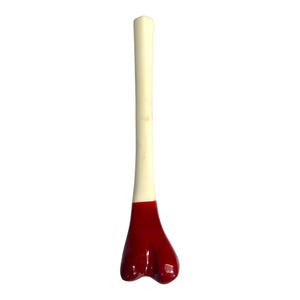 Puede incluir: Un tapón de botella con forma de hueso, con un tallo color crema y una base roja brillante. La base tiene un diseño redondeado de tres lóbulos. El tapón está diseñado para encajar en la abertura de una botella.