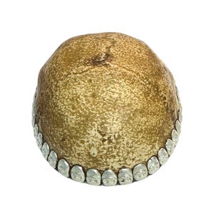 May include: A round, textured, beige-colored decorative object with a silver-colored border. The border is adorned with small, repeating, skull-shaped designs. The object has a rough, organic appearance.