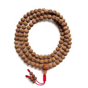 Peut inclure: Un chapelet de perles de prière marron avec un pompon rouge. Les perles sont rondes et texturées, et le pompon est fait de fil rouge. Les perles sont disposées en cercle sur fond blanc.