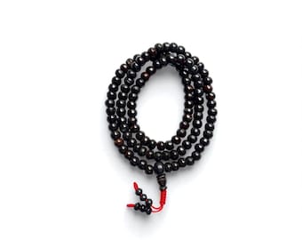Puede incluir: Un collar de cuentas marrón oscuro con una borla roja. Las cuentas son redondas y parecen ser de madera. El collar está dispuesto en un patrón circular sobre un fondo blanco.