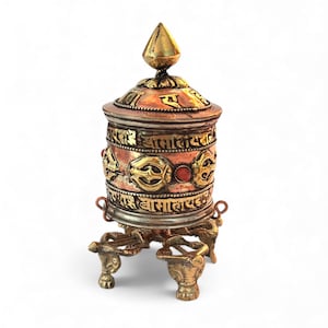 May include: A copper and brass prayer wheel with intricate carvings and a conical top. The wheel features embossed patterns, a red stone accent, and is supported by a decorative three-legged base. The prayer wheel is a religious item.