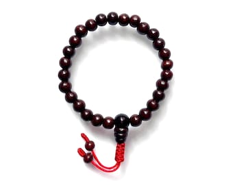 Peut inclure: Un bracelet de perles en bois brun foncé avec un pompon rouge. Le bracelet est composé de petites perles rondes enfilées ensemble. Le pompon est attaché au bracelet avec un nœud et comporte deux petites perles à l'extrémité. Le bracelet est sur fond blanc.