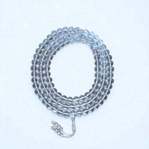 Puede incluir: Un collar de cuentas transparentes y grises, probablemente piedras preciosas, dispuestas en un patrón circular. Las cuentas están ensartadas, con una borla decorativa plateada en el extremo. El fondo es blanco liso, resaltando las cuentas.