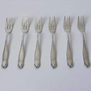 Pode incluir: Seis garfos prateados com um design clássico dispostos sobre uma superfície branca. Cada garfo tem três dentes e um cabo com um padrão floral decorativo na base. Os garfos são feitos de metal brilhante e refletor.