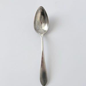 Może przedstawiać: Srebrna łyżka z owalną miseczką i długim, smukłym trzonkiem. Łyżka ma błyszczącą, odblaskową powierzchnię i wydaje się być wykonana z metalu. Trzonek ma grawerowany wzór w pobliżu końca.