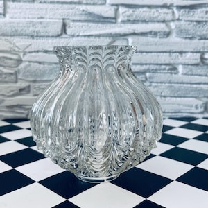 Könnte beinhalten: Eine klare Glasvase mit einem strukturierten, gerippten Design. Die Vase hat einen breiten, abgerundeten Körper und eine gerüschte Oberseite. Die Vase steht auf einer schwarz-weiß karierten Oberfläche, mit einer weißen Backsteinmauer im Hintergrund.