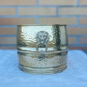 Könnte beinhalten: Ein goldfarbener, gehämmerter Metalltopf mit einem Löwenkopf-Design auf der Vorderseite. Der Topf ist tonnenförmig und hat eine gehämmerte Textur.