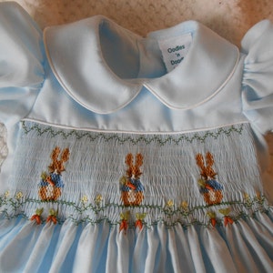 May include: A light blue dress with a Peter Pan collar and short puffed sleeves. The bodice features white smocking with embroidered brown rabbits wearing blue jackets and holding orange carrots, along with small yellow flowers and green vines.