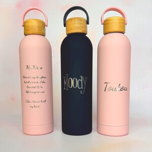 Three personalised insulated water bottles with engraved names and message text, shown in blush pink and black with bamboo-effect lids. Custom reusable bottles for personal gifts, branded giveaways or company merchandise.
