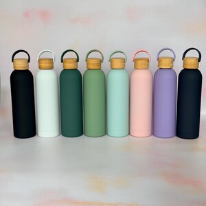 Row of reusable metal water bottles in black, white, green, mint, pink and purple, each with a wooden lid and carry loop, shown on a clean neutral background.