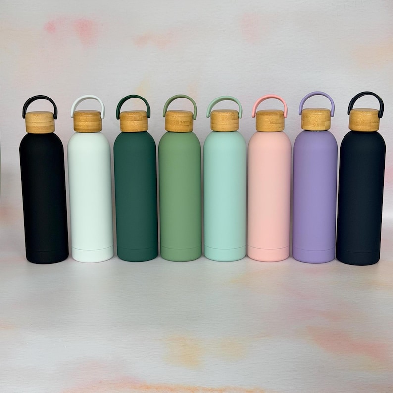 Row of reusable metal water bottles in black, white, green, mint, pink and purple, each with a wooden lid and carry loop, shown on a clean neutral background.