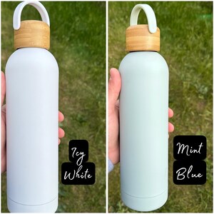 Side-by-side image of two rubber-coated water bottles held outdoors against green grass. The left bottle is a soft Icy White shade with a bamboo lid and matching white loop handle. The right bottle is a cool Mint Blue with the same bamboo lid and matching mint loop handle.
