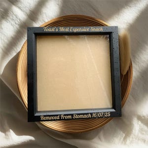 May include: A black picture frame with gold text reading "Toast's Most Expensive Snack" and "Removed From Stomach 16/07/25". The frame is empty and rests on a woven, circular tray.