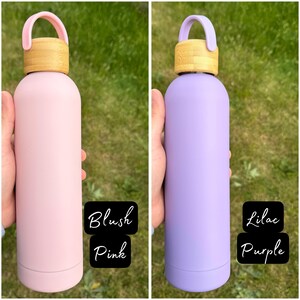 Two pastel-coloured water bottles held against a grassy background. The left bottle is Blush Pink with a bamboo lid and pink loop handle. The right bottle is Lilac Purple with the same bamboo lid and a matching lilac handle.