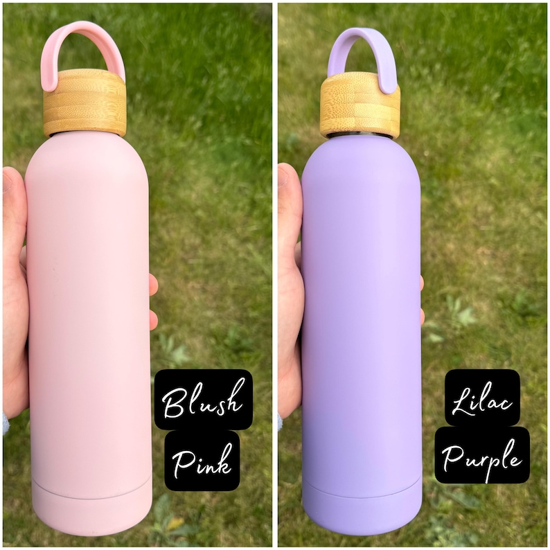 Two pastel-coloured water bottles held against a grassy background. The left bottle is Blush Pink with a bamboo lid and pink loop handle. The right bottle is Lilac Purple with the same bamboo lid and a matching lilac handle.