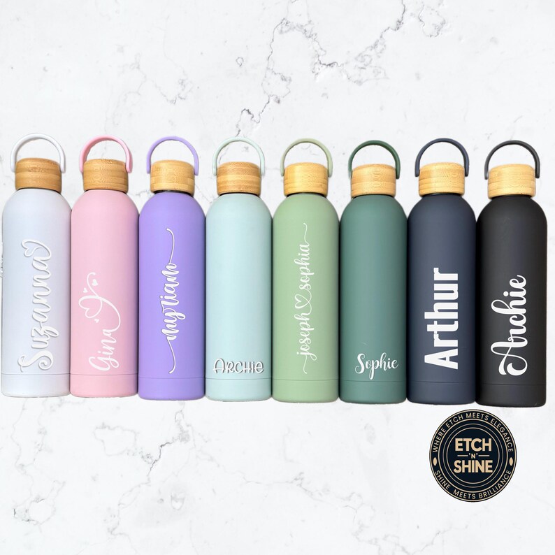 Row of colourful personalised stainless-steel water bottles with engraved names in various fonts, bamboo lids and matte finish, reusable eco drinkware by Etch ’n’ Shine.