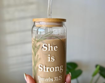 Puede incluir: Un vaso de vidrio transparente con tapa de bambú y pajita de vidrio, lleno de una bebida de café. El vaso tiene las palabras "She is Strong Proverbs 31:25" en letras blancas, con un diseño de hoja verde.
