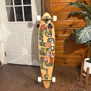 Könnte beinhalten: Ein Longboard-Skateboard mit einem floralen und Schlangen-Design, das auf das Deck gemalt ist. Die Schlange ist schwarz und grün mit einer rosa Zunge. Die Blumen sind rosa, orange und gelb. Die Räder sind weiß und die Achsen sind golden.