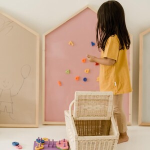 以下が含まれることがあります： ピンクの磁石ボードに、カラフルな磁石の文字と形を使って遊んでいる子供。蓋付きの籐のバスケットが、床に置かれた木製の形合わせパズルと木製のペグパズルの隣に置かれている。