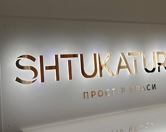 May include: A backlit sign with the word "SHTUKATURKA" in gold lettering. Below, in smaller gold letters, is the text "ПРОСТІР КРАСИ". The sign is mounted on a white surface.
