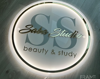 May include: A white circular sign with a gold trim and a gold inscription that reads "Saba Studio beauty & study". The letters "S" are large and gray, with the inscription in front of them.