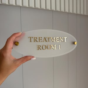 May include: An oval-shaped, frosted acrylic sign featuring the gold-coloured text "TREATMENT ROOM 1" in a serif font. The sign is secured with two small, circular gold-toned metal standoffs on the left and right sides of the oval plate.