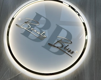 May include: A round, illuminated sign with the words "Beauty Bliss" in elegant, gold-colored script. The sign has a white face with a gold trim and a subtle, glowing light around the edge. The background is a light gray wood.