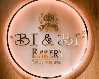 May include: A circular, illuminated wall sign featuring the metallic text BI & BI BAKERY and the Cyrillic phrase ТАМ ДЕ ЖИВЕ ДУША. Above the text is a graphic of a cupcake with wheat stalks. The sign has a warm, glowing backlight effect.