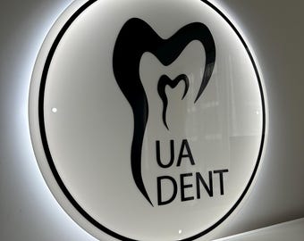 May include: A round, illuminated sign with a white face and a black border. The sign features a stylized tooth graphic and the text "UA DENT" in black. The sign is mounted on a wall.