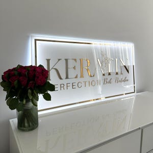 Może przedstawiać: Prostokątny szyld ze złotym napisem "KERATIN" i "PERFECTION Bik Natalia" poniżej. Szyld jest biały ze złotą obwódką i podświetlony. Bukiet czerwonych róż w szklanym wazonie po lewej stronie.